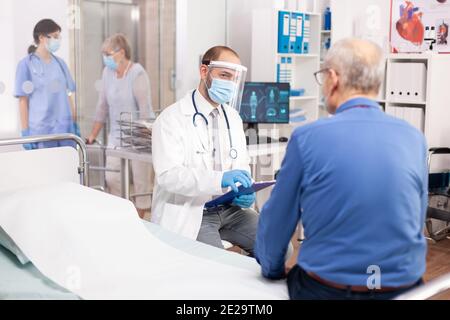 Medical staff questioning senior man in clinic consultation room wearing visor in the course of covid19. Physician practitioner in the course of private modern clinic appointment during coronavirus pandemic. Stock Photo