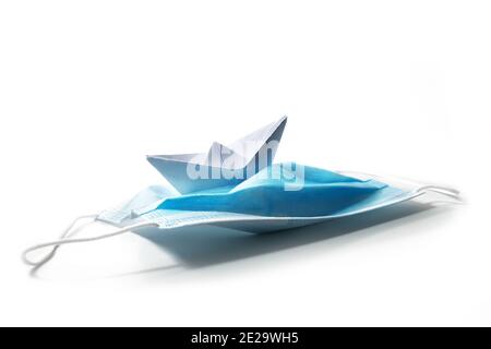 Paper boat keeping course on a surgical face mask against covid-19 pandemic as a ship riding in stormy seas, coronavirus crisis concept, isolated on w Stock Photo