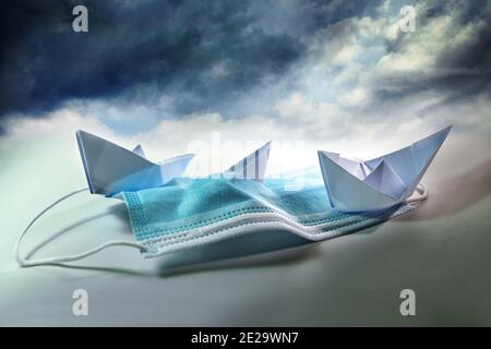 Three paper boats struggle against sinking on a surgical face mask under a stormy sky, metaphor for perseverance in coronavirus times, covid-19 pandem Stock Photo