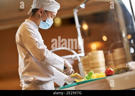 Side-view photo of a male chef wearing protective mask while chopping vegetables on a cutting board Stock Photo