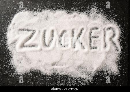 German word Zucker, meaning sugar, written in spilled out sugar crystals on a dark slate, high angle view from above, selected focus Stock Photo