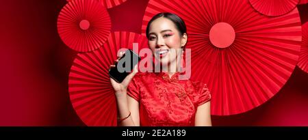 Beautiful Asian woman wearing traditional Chinese qipao dress holding mobile phone with empty screen in oriental style red banner background Stock Photo