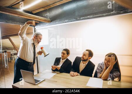 Mad man. Angry boss with megaphone screaming at employees in office, scared and annoyed colleagues listening at the table stressed out. Funny meeting, business, office concept. Screaming mad. Stock Photo