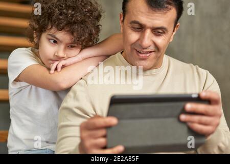 Middle aged latin father using tablet pc while spending time together with his son, sitting on the stairs at home. Family, children, technology Stock Photo