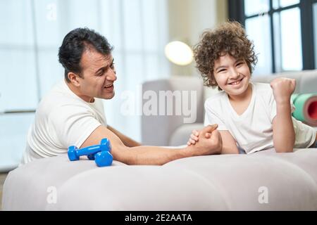 Happy latin boy looking satisfied after armwrestling together with his father at home, lying on the couch. Healthy lifestyle, family concept Stock Photo