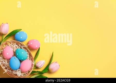 Easter greeting card invitation. Multi-colored painted eggs in a basket, pink tulips on a yellow background. Stock Photo