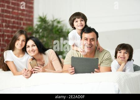 Portrait of happy latin family with cute kids smiling at camera while using digital devices, lying on the bed together. Family, parenthood, technology concept Stock Photo
