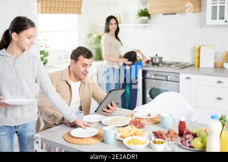 Happy latin family having dinner at home. Middle aged husband using tablet pc, watching news while his daughter and wife serving table, standing in the kitchen Stock Photo