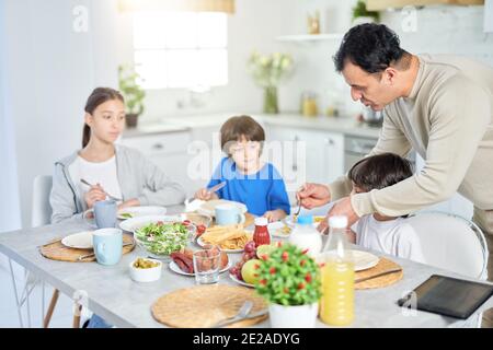 Caring latin father serving his adorable kids while having breakfast together at home. Childhood, parenthood, latin cuisine concept Stock Photo