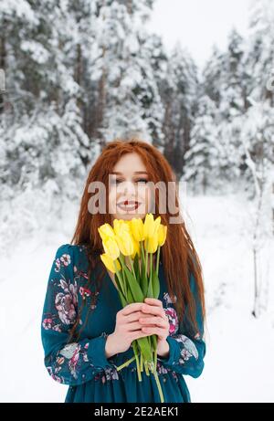 A girl with red hair holds a bouquet of yellow tulips on a winter background. Flowers as a gift for women's Day. March 8.A beautiful girl smiles. Stock Photo