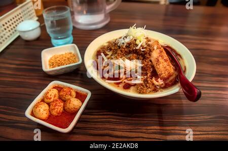 A bowl of tantanmen noodles with sides of fried garlic bits and spicy marinated boiled quail eggs on a wooden table in a restaurant Stock Photo