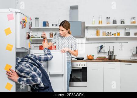 young woman giving screwdriver to handyman repairing fridge in kitchen Stock Photo