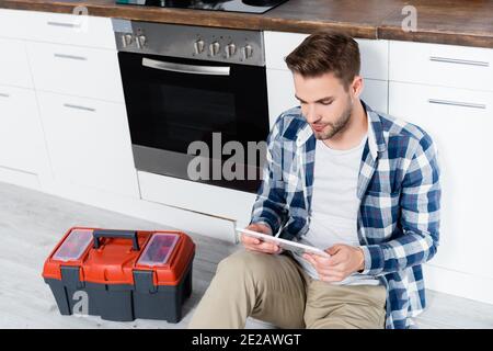 young man looking at tablet while sitting near toolbox and oven on floor in kitchen Stock Photo