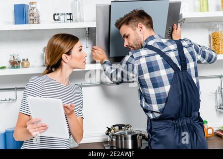 young handyman looking at tablet while repairing extractor fan near woman in kitchen Stock Photo