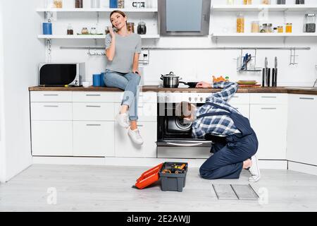 full length of smiling woman talking on smartphone while sitting on table near handyman repairing oven in kitchen Stock Photo