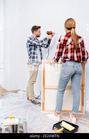 full length of young woman holding frame near man drilling wall at home Stock Photo