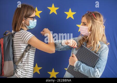 Elbow bump greeting by two female students wearing protective masks with EU flag in the background. Stock Photo