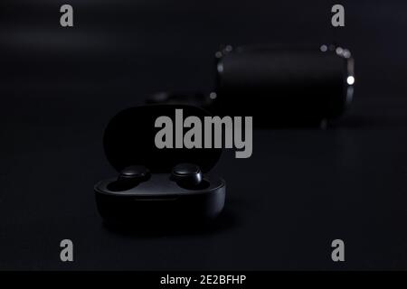 Black wireless headphones in charging case on black with wireless speaker on background. Stock Photo