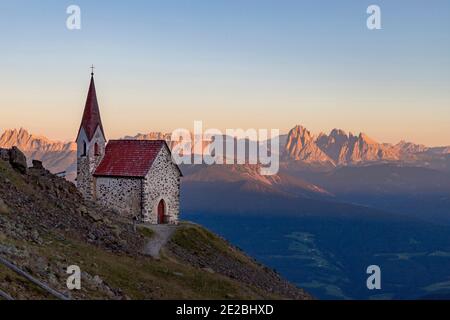 Little chapel and view over orange-lit Dolomite mountain range at sunset in summer, South Tyrol / Alto Adige, Italy Stock Photo