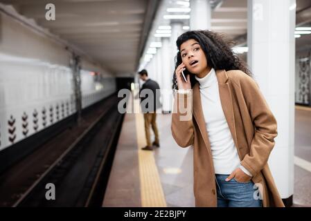 surprised african american woman talking on smartphone and looking away in subway on blurred background Stock Photo