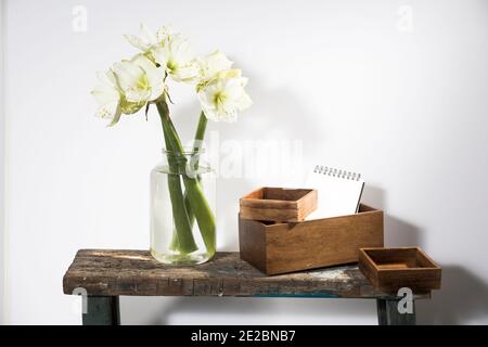 Bouquet of three white lilies in a tall glass vase on a beige vintage bench against a gray wall. Wodden boxes different size with scetchbook. Copy spa Stock Photo