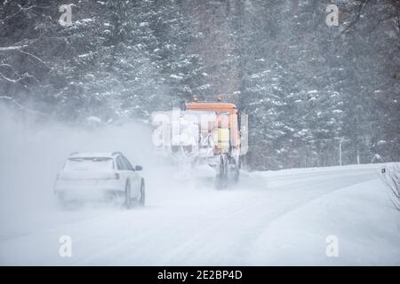 Left-handed driving road maintenance truck clears snow from the road in the forest with a car driving behind it. Stock Photo