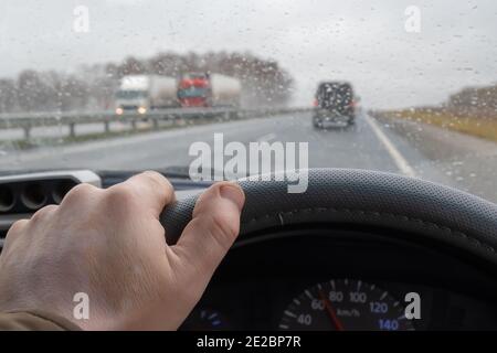 the driver's hand on the steering wheel of a car that is driving on the highway among other cars and trucks in rainy cloudy weather Stock Photo