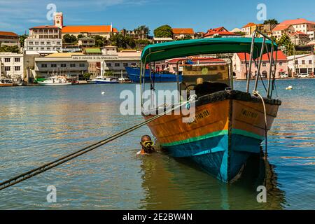 The excursion boat "Family Thing 108" lies at anchor in the Carenage, the horseshoe-shaped harbour of St. George's, Grenada. The name Carenage comes from the French word for cleaning and repairing ship hulls. A man wearing diving goggles swims beside its hull. Across the water stands the warehouse of the trading company Geo. F. Huggins & Co. Stock Photo