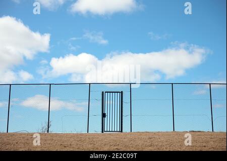 Low angle of chain link fence with door on dry grassy terrain against cloudy blue sky Stock Photo