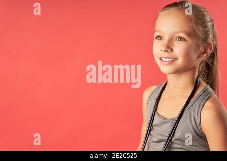 Cute joyful girl with skipping rope around her neck looking away and smiling Stock Photo
