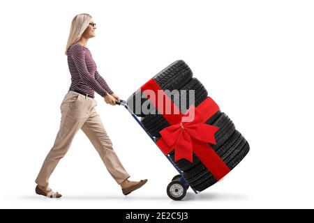 Full length profile shot of a young woman pushing a hand-truck loaded with tires tied with a red ribbon isolated on white background Stock Photo