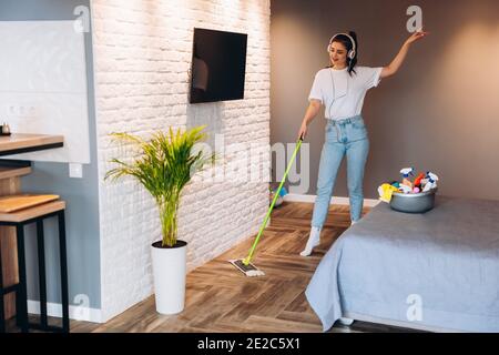 Full length photo of happy cheerful woman is washing floor in room with mop having fun. Imagine of housewife dancing in headphones while cleaning indo Stock Photo