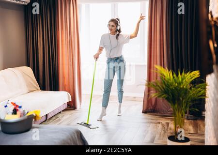 Full length photo of happy cheerful woman is washing floor in room with mop having fun. Imagine of housewife dancing in headphones while cleaning indo Stock Photo