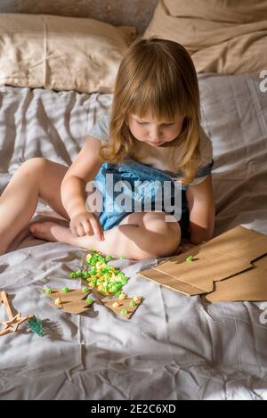 small kid making Christmas crafts, family celebration.girl makes a Christmas tree on the bed. Scissors and cardboard. Zero Waste Stock Photo