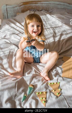 kids making Christmas crafts, family celebration.girl makes a Christmas tree on the bed. Scissors and cardboard. Zero Waste Stock Photo