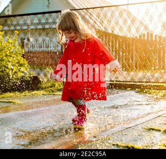Cute girl in a red jacket is jumping in the puddle.The setting warm summer or autumn sun. summer in the village. Stock Photo