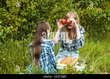 Happy smiling woman and fun enjoying kid, mother holding the red apples near the eyes.Picnic in the park, happy family. Sunny summer day outdoors Stock Photo