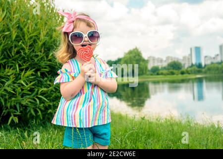 Fashionable baby girl, sunglasses, lollipop like a heart.little three year old girl licks a colored swirling candy on a walk in the park summer day Stock Photo