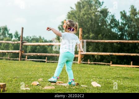 Little blond girl climbing over the wooden fence, summer day on the forest background. Vocation in the village. Life Style Outdoors Stock Photo