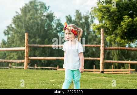 Little blond girl climbing over the wooden fence, summer day on the forest background. Vocation in the village. Life Style Outdoors Stock Photo