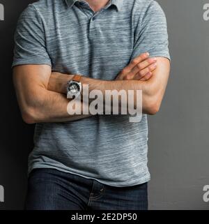 Young man in a gray shirt wearing a watch with his arms cross leaning on a wall Stock Photo