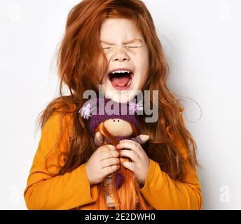 Cheerful small red haired girl in yellow comfortable longsleeve standing holding toy doll in hands and screaming feeling excited Stock Photo