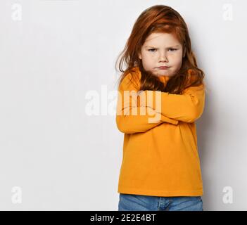 Serious small red haired girl in yellow comfortable longsleeve standing with crossed hands and frowning Stock Photo