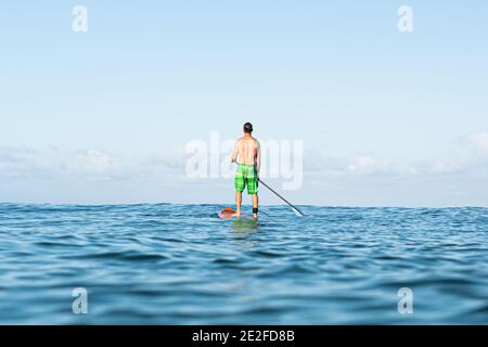 A young man in shorts stands on a paddle board at sea waiting for the next wave, blue sky. Stock Photo