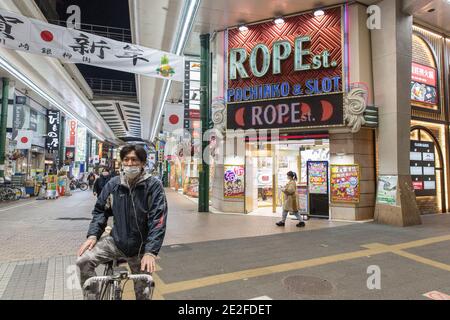 Kawasaki, Japan. 13th Jan, 2021. A man wearing a facemask on a bicycle in front of a Pachinko & Slot venue in Ginryugai street in Kawasaki. Credit: SOPA Images Limited/Alamy Live News Stock Photo