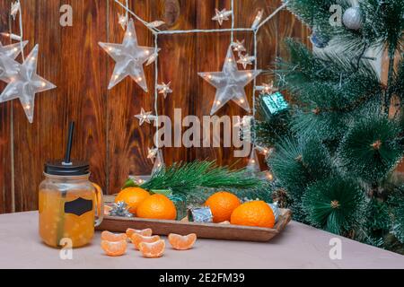 Mandarins and fruit wedges on New Year's table. Christmas tree and jar of fresh juice with handle. Free label for your text. Blurred background with g Stock Photo
