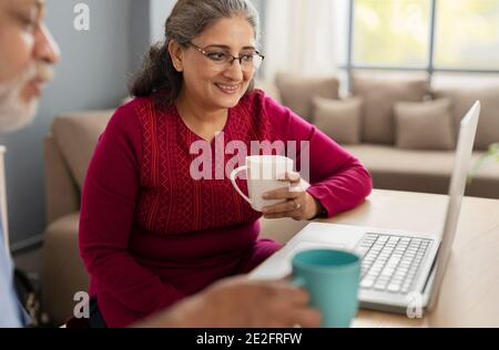 A SENIOR ADULT WOMAN HOLDING MUG LOOKING AT LAPTOP WITH HUSBAND BESIDES Stock Photo