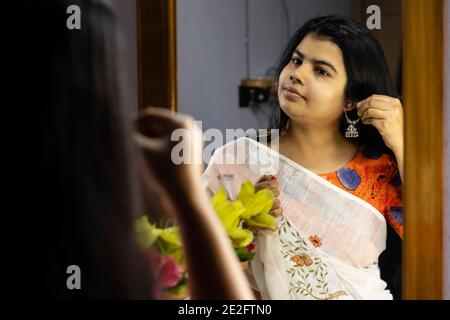 a beautiful Indian woman in white saree wearing silver earrings in front of mirror with smiling face Stock Photo