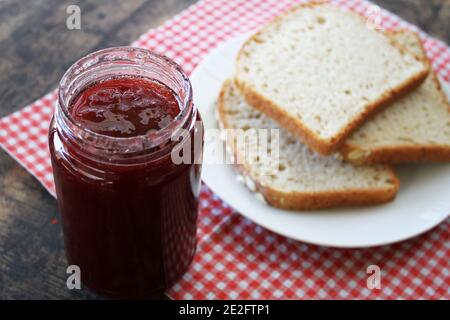 A jar of strawberry jam next to three slices of bread against a rustic table backdrop. Stock Photo