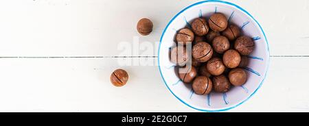 Organic Macadamia nuts (Macadamia integrifolia) in shell on wooden white table. Top view, overhead,  banner Stock Photo